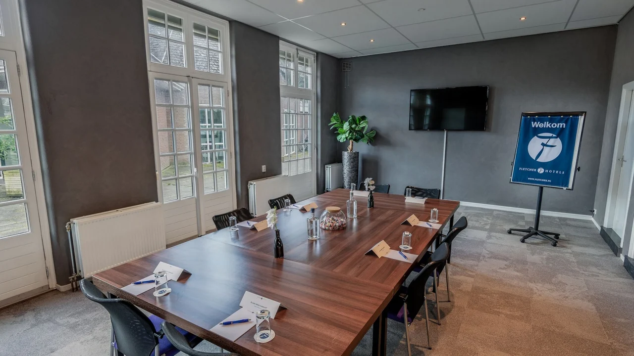 Lange vergadertafel in de orangerie met hoge ramen en televisie