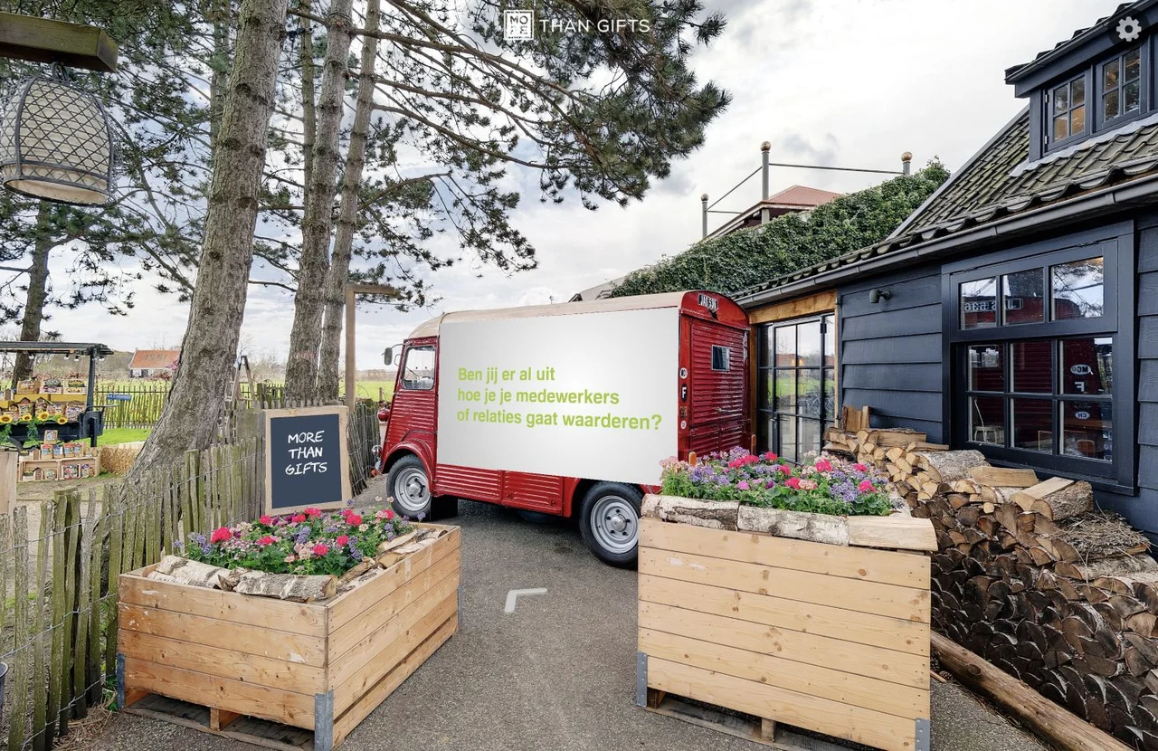 Entree van de virtuele lentemarkt van More Than Gifts met een rode bestelwagen, bloemenbakken en een houten gebouw